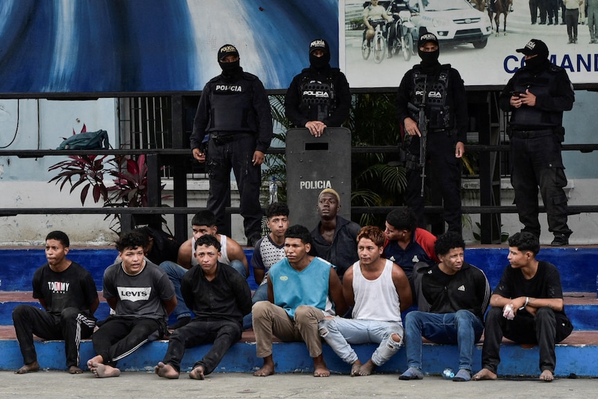 Four police officers stand behind twelve men seated on a set of stairs.