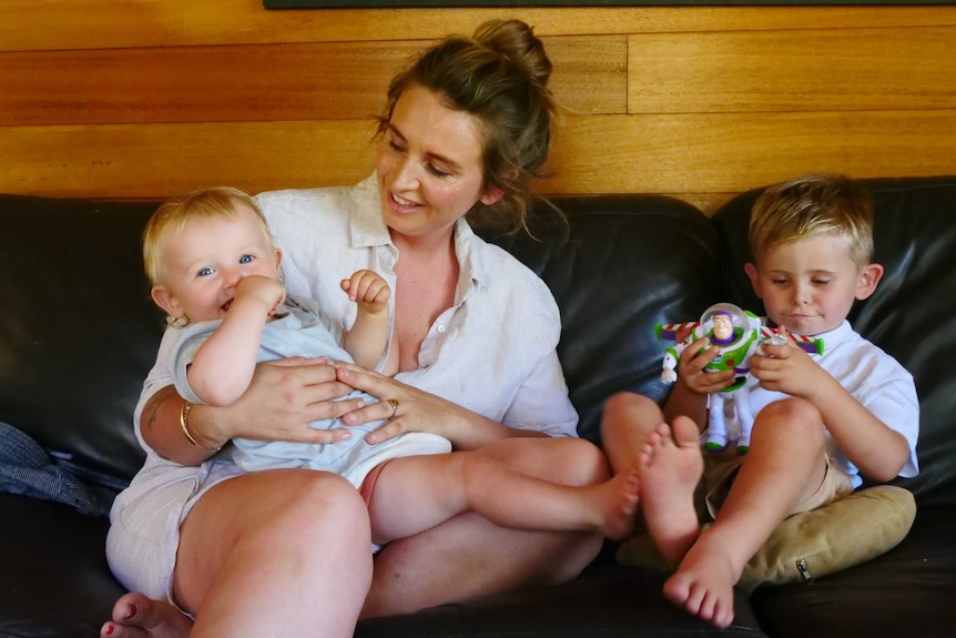 Mother holding her baby on couch with toddler playing toys sitting beside her on the couch.