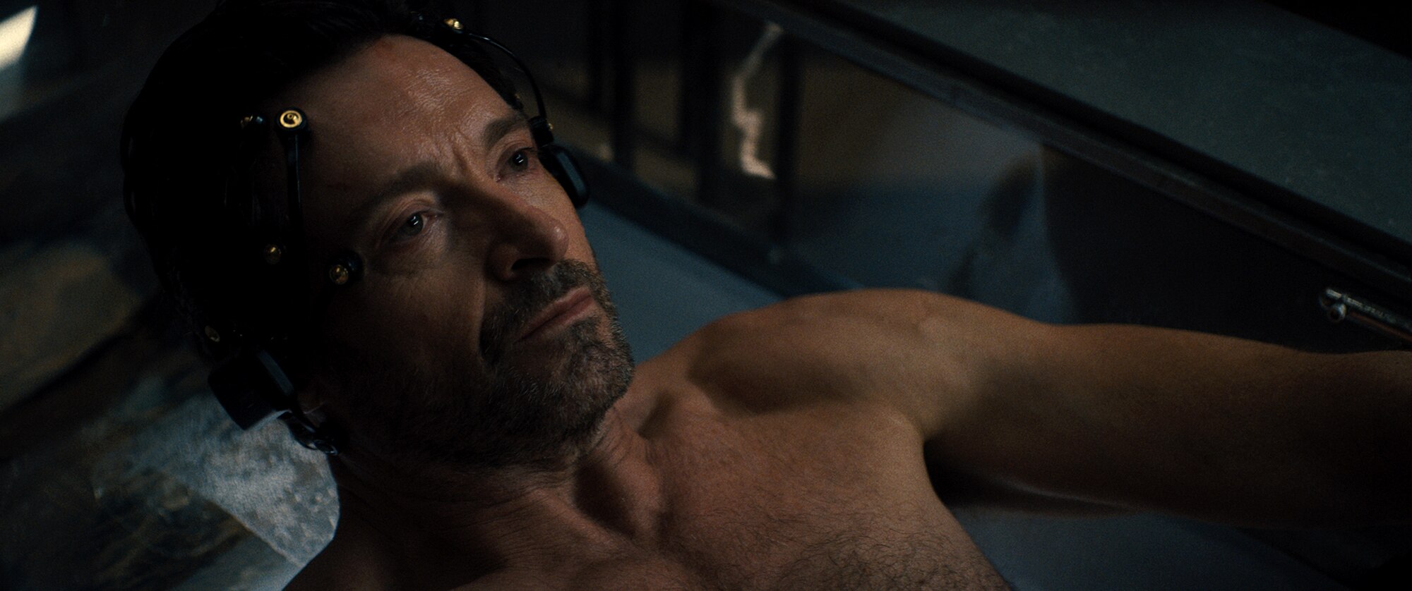 A close up on a dark and brooding Hugh Jackman. He is lying down in a high-tech pod-like machine and is intently focused.