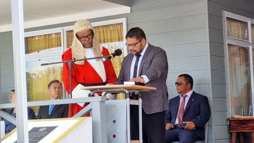 Man at podium looks down as another man in red robes and solicitor's wig stands by him.