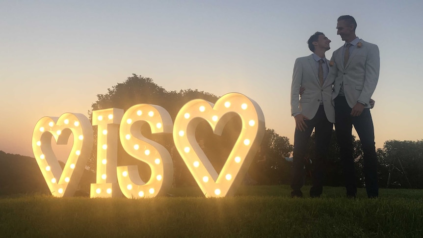 Craig Burns with his partner Luke Sullivan next to an illuminated "love is love" sign.