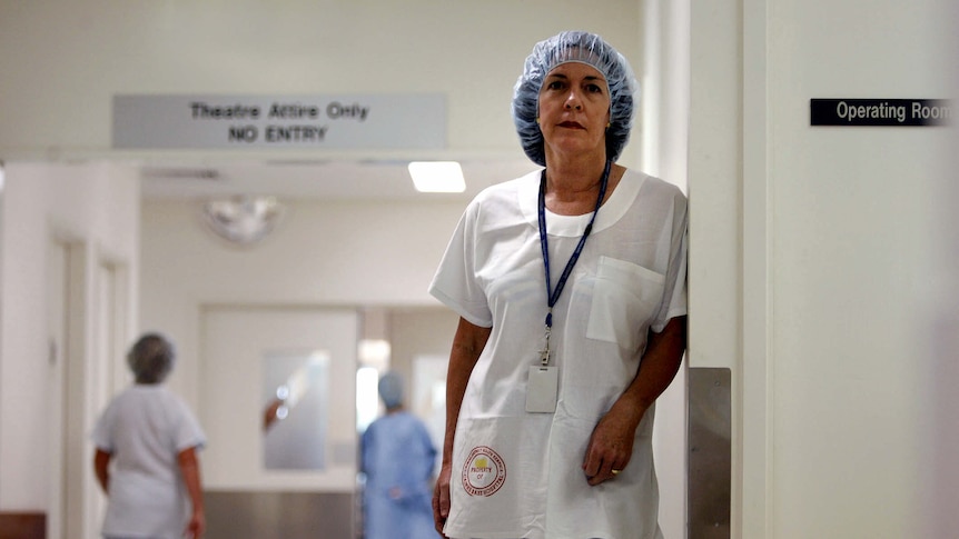 Caroline de Costa, dressed in full hospital scrubs, stands leaning against a wall in a hospital corridor.