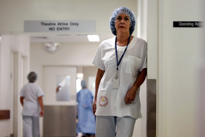 Caroline de Costa, dressed in full hospital scrubs, stands leaning against a wall in a hospital corridor.