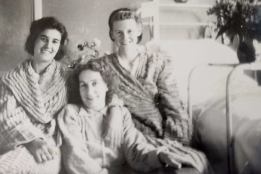 Black and white photo of three women sitting in front of a hospital bed.