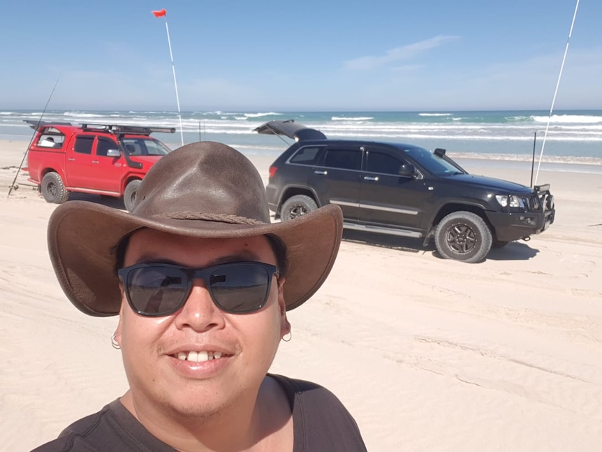 Harrison taking a selfie with some four-wheel drives on a beach, wearing a hat and sunnies.