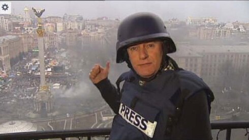 Philip Williams stands on a balcony in a bulletproof vest that has "Press" on the front and a helmet, pointing to smoke below.
