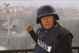 Philip Williams stands on a balcony in a bulletproof vest that has "Press" on the front and a helmet, pointing to smoke below.