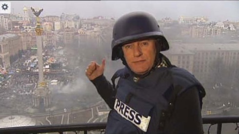 A man stands on a balcony in a bulletproof vest that has "Press" on the front and a helmet, and pointing to smoke below.