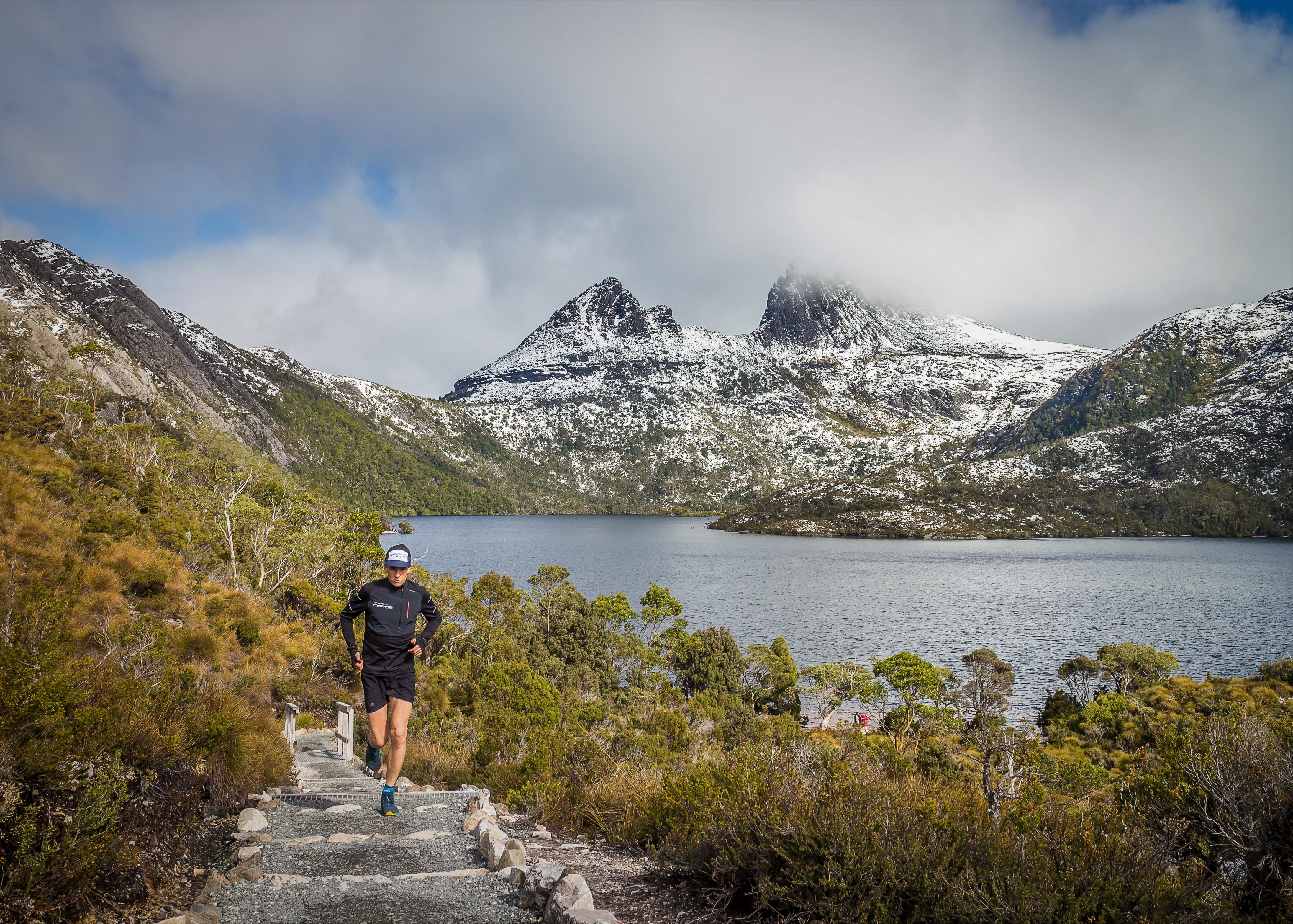 A runner running along a trail towards the camera, with a snow dusted Cradle Mountain behind.