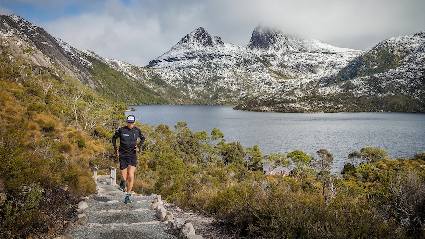 A runner running along a trail towards the camera, with a snow dusted Cradle Mountain behind.