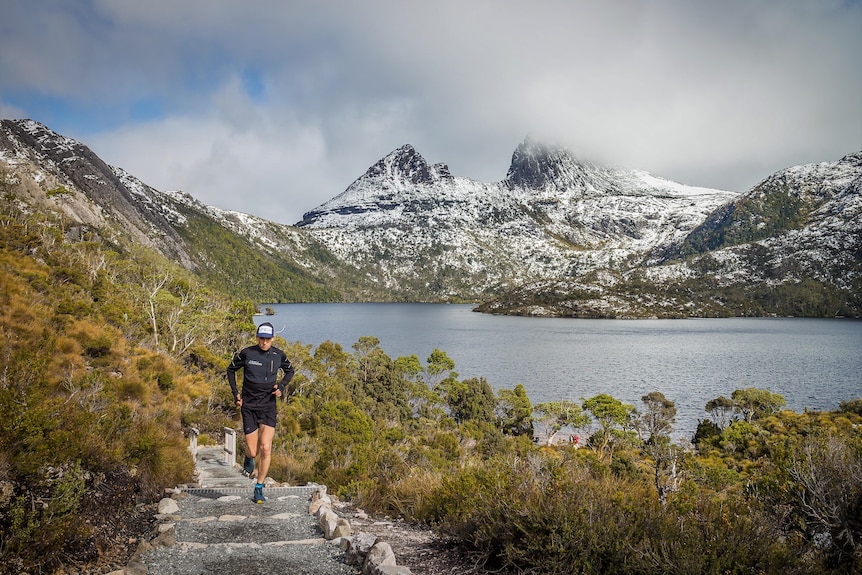 A runner running along a trail towards the camera, with a snow dusted Cradle Mountain behind.