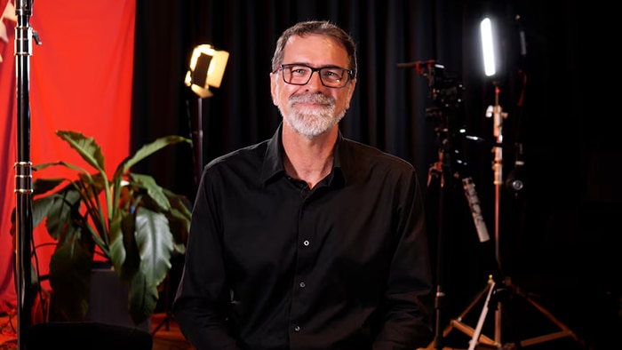 Man smiling standing in front of film lights, a plant and a red and black backdrop.