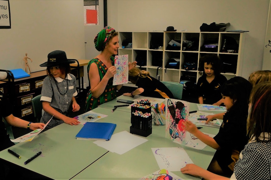 Woman with bohemian head dress holds up abstract drawing to students sitting at table. 