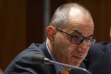 Home Affairs Secretary Mike Pezzullo speaking at a committee hearing, wearing glasses and a yellow tie. Mitch Fifield watches on