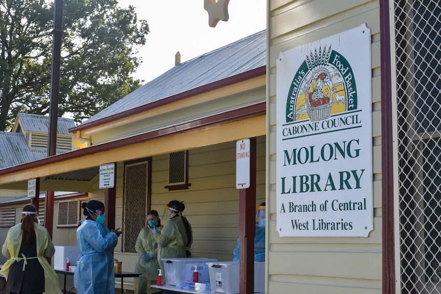 A sign reads 'Molong Library' and nursing staff in full PPE stand around boxes of COVID-19 swabs.