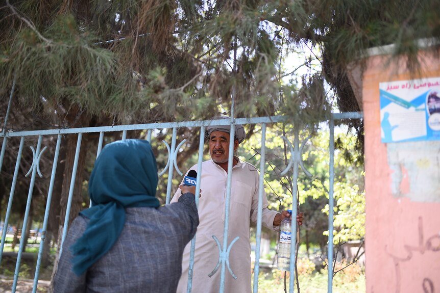 A woman wearing a hijab puts her microphone through a fence to interview a man wearing a white tunic.