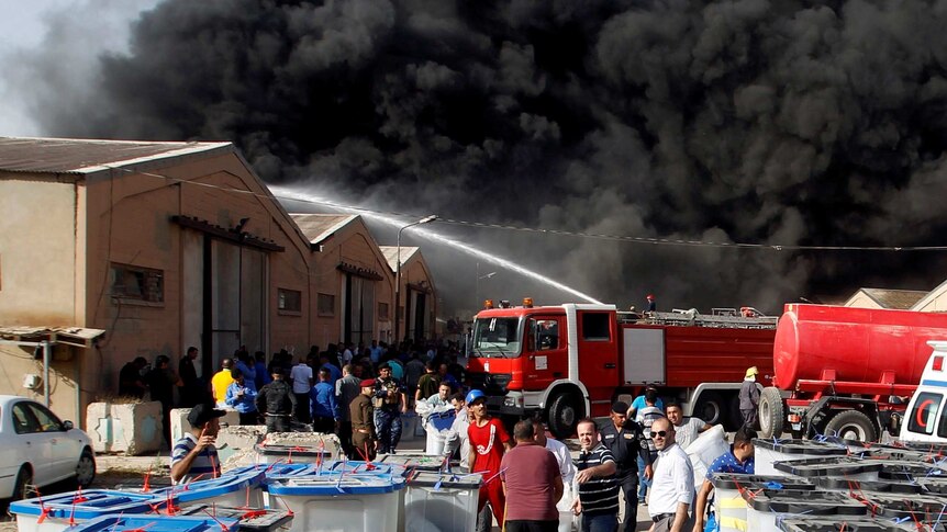 Firefighters battle a fire at a warehouse site with thick black smoke rising in the background.