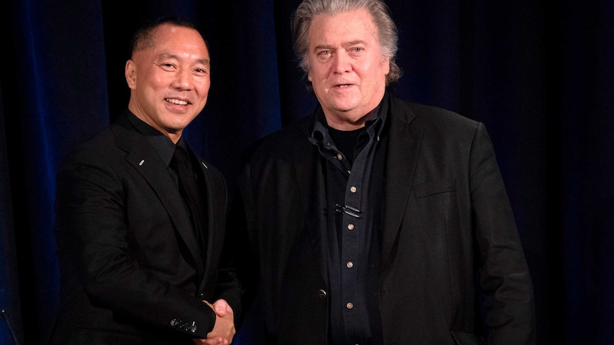 Two men dressed in black clasp hands in front of a dark blue curtain.