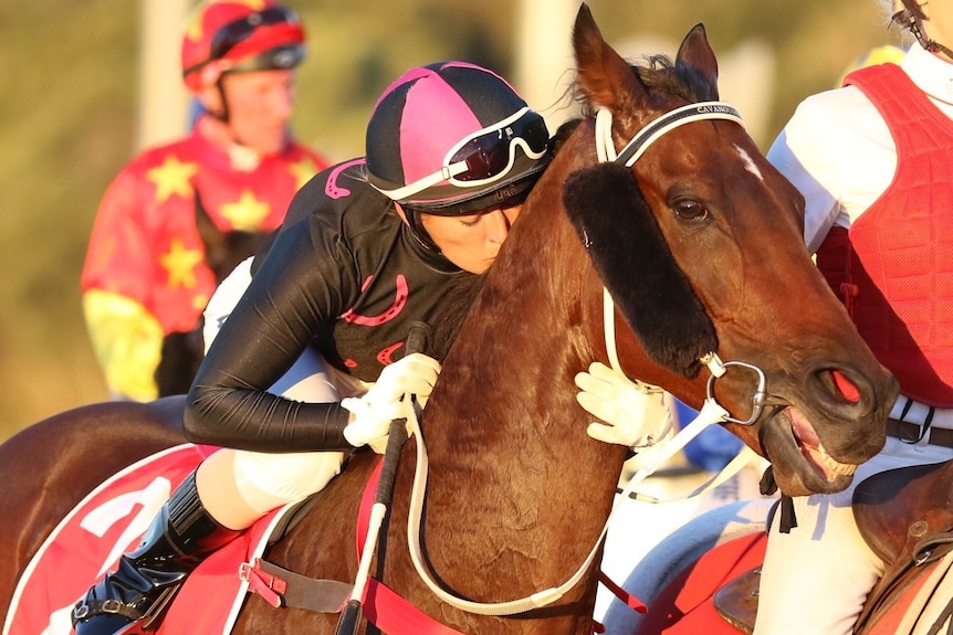 Skye Bogenhuber leans forward and kisses a horse on the back of the neck whilst sitting on the horse wearing black and pink silk