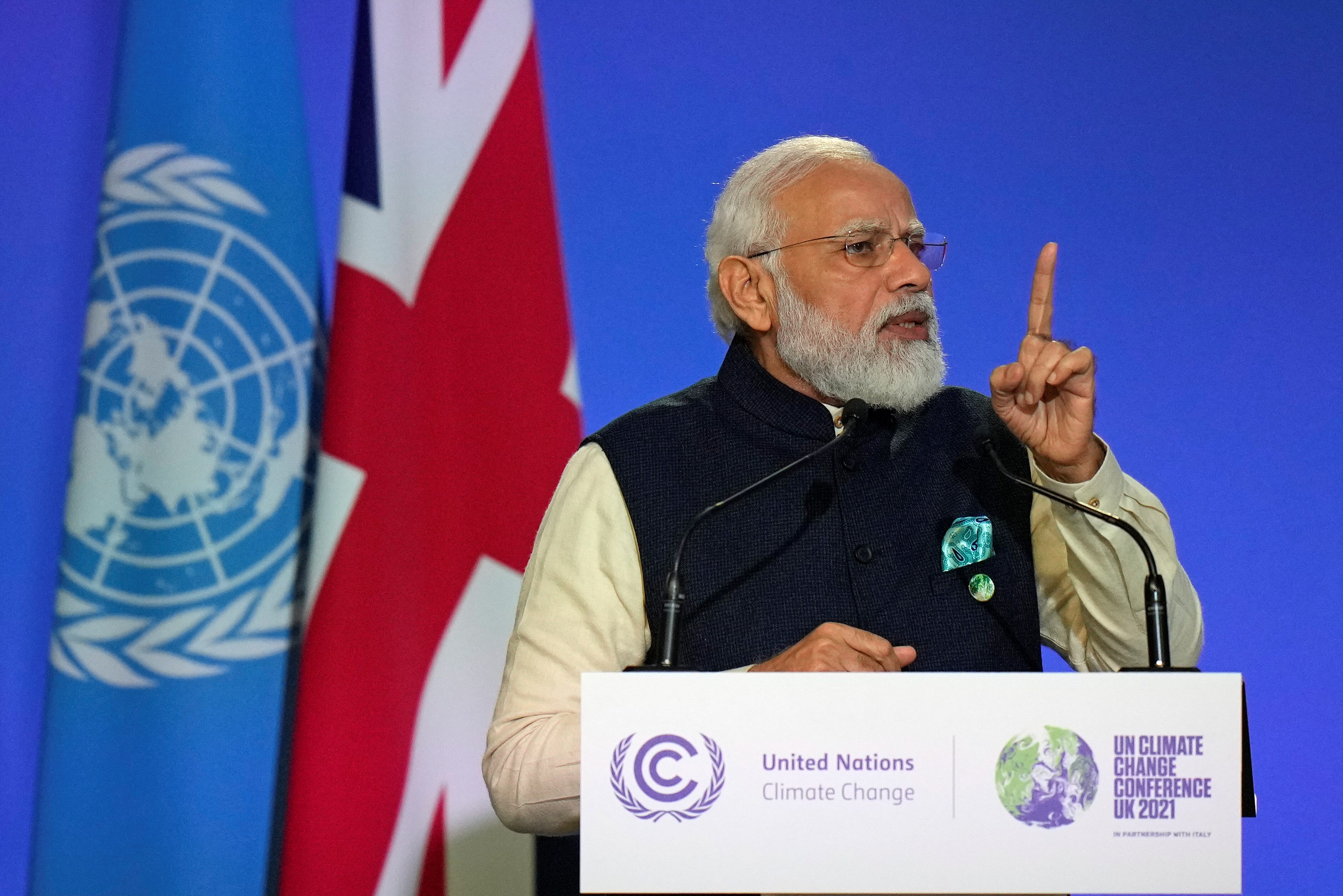 Man in vest wearing glasses points as he gives a speech in front of two flags in Glasgow.