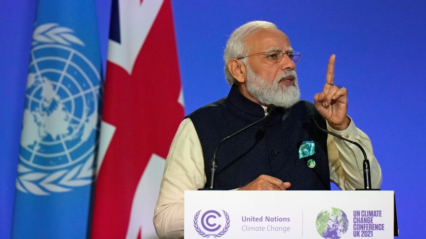 Man in vest wearing glasses points as he gives a speech in front of two flags in Glasgow.