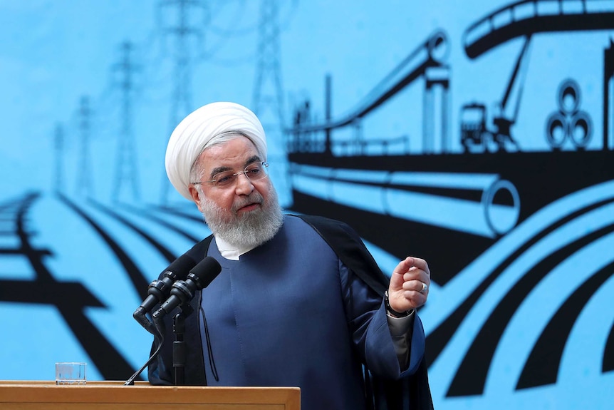 President Hassan Rouhani wears a white turban and speaks at a lectern in front of a blue backdrop.