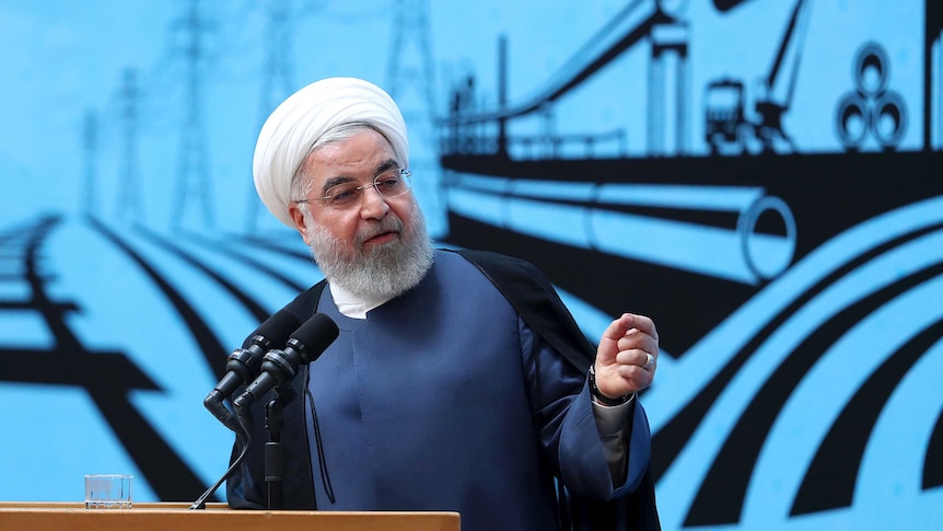 President Hassan Rouhani wears a white turban and speaks at a lectern in front of a blue backdrop.