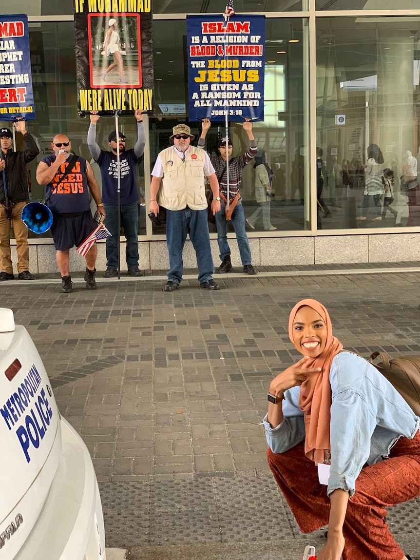 A woman wearing a hijab is smiling while holding a peace sign in front of a group of men with anti-Muslim posters.