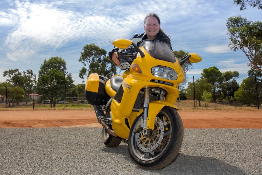 A woman holding a motorcycle helmet and leaning forward over a yellow motorbike