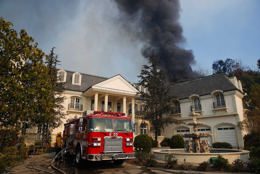 A fire truck is parked outside a mansion in Bel-Air, there is smoke behind it from a wildfire.