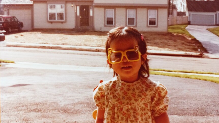 A child with dark hair is seen wearing iconic yellow sunglasses and a floral dress in a suburban street in 1980s Virginia. 