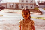 A child with dark hair is seen wearing iconic yellow sunglasses and a floral dress in a suburban street in 1980s Virginia. 