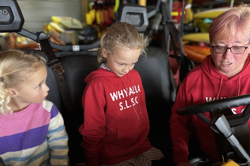 Two young blonde girls sit in a buggy beside a woman with short blonde hair and glasses.