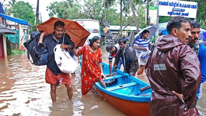 People in India wade in flooded waters. One woman is climbing into a boat and a man is carrying an umbrella.