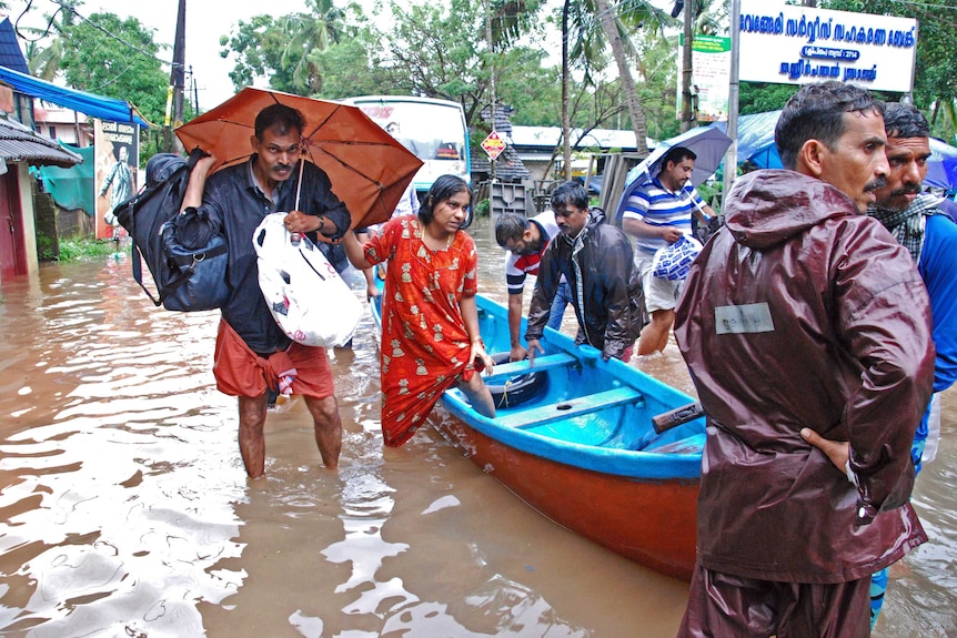 People in India wade in flooded waters. One woman is climbing into a boat and a man is carrying an umbrella.