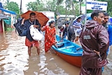 People in India wade in flooded waters. One woman is climbing into a boat and a man is carrying an umbrella.