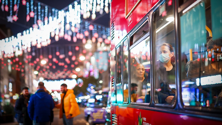 A woman in a face mask looks out the window of a red double decker bus at Christmas lights 