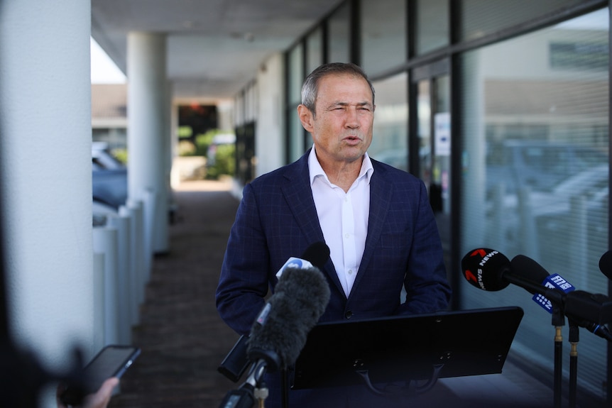 A wide shot of WA Premier Roger Cook standing and speaking in front of reporters' microphones outside an early voting centre.