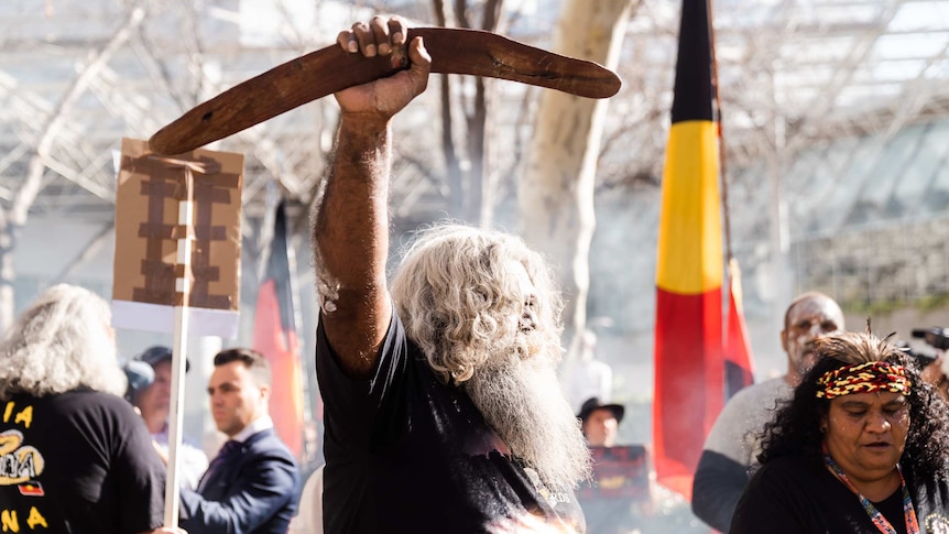 A man holds a boomerang in the air, an Aboriginal flag behind him, a woman wears a red, yellow and black headband with feathers.