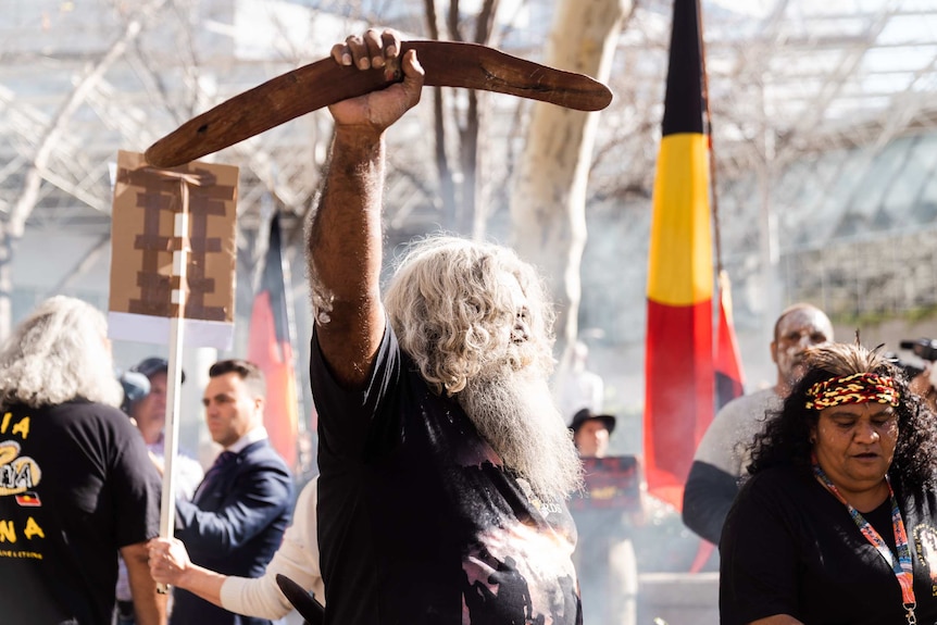 A man holds a boomerang in the air, an Aboriginal flag behind him, a woman wears a red, yellow and black headband with feathers.