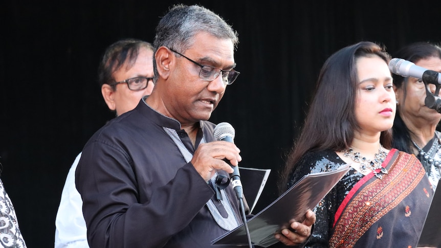Man in black shirt and glasses holds microphone and reads from paper he is holding