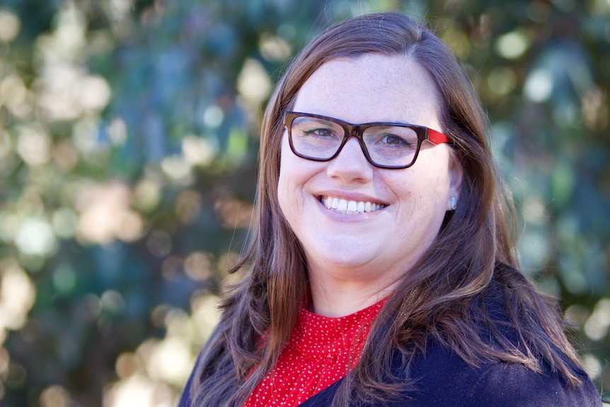 A woman with glasses smiles at the camera, trees in background.