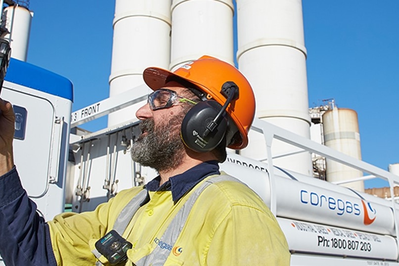 A man wearing a hard hat looks at a device standing in front of series of gas tanks.