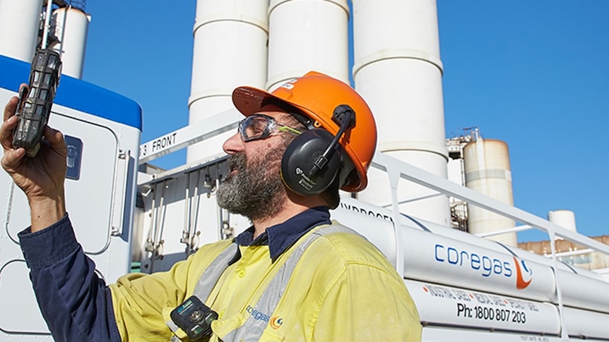 A man in high-vis clothing, a hard hat and ear protectors monitors equipment while standing in front of a Coregas plant.