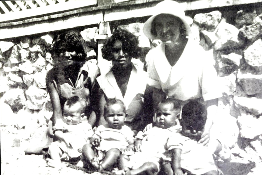 A nun sits on the ground with two woman and four babies sat in front of them.