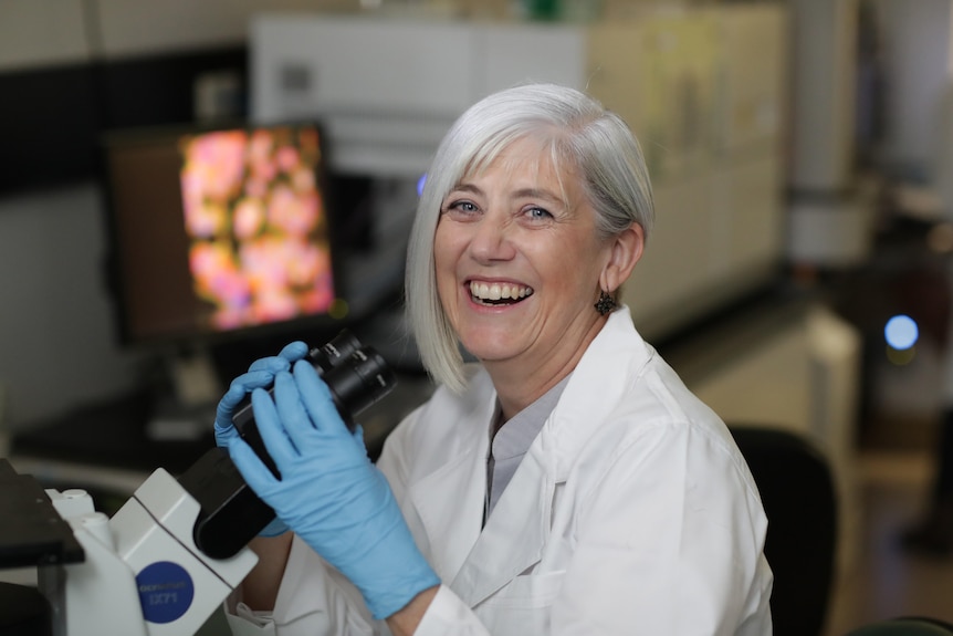 A woman with a lab coat and gloves smiles while holding a microscope.