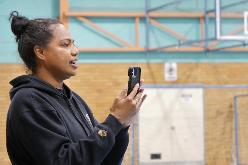 A woman is holding a phone and watching action off camera on a basketball court.