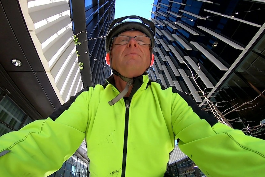 Close-up of a man in a bright yellow cycling jacket, and a helmet, riding between two city buildings.