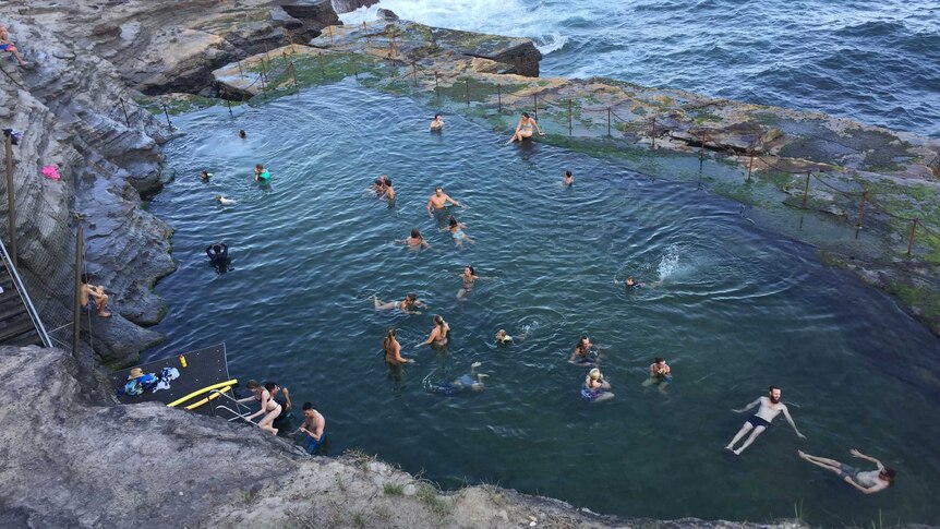People swim and frolic in the waters of an unusually-shaped ocean pool at the bottom of a cliff face next to the ocean