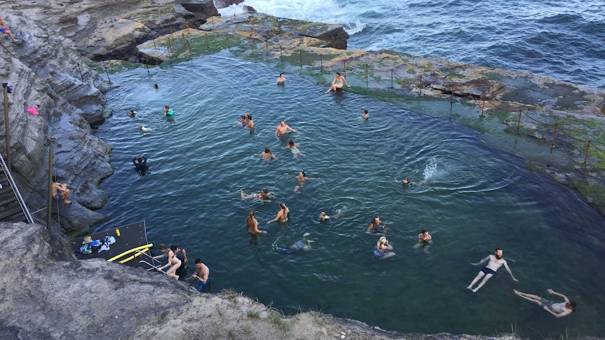 People swim and frolic in the waters of an unusually-shaped ocean pool at the bottom of a cliff face next to the ocean